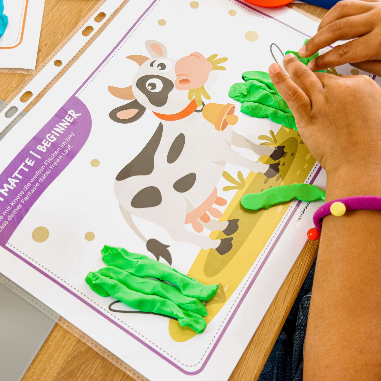 Kind modelliert mit grüner Knete das Gras auf einer mykidslounge Beginner Knetmatte mit einer fröhlichen Kuh – ideal für erste Kneterfahrungen.