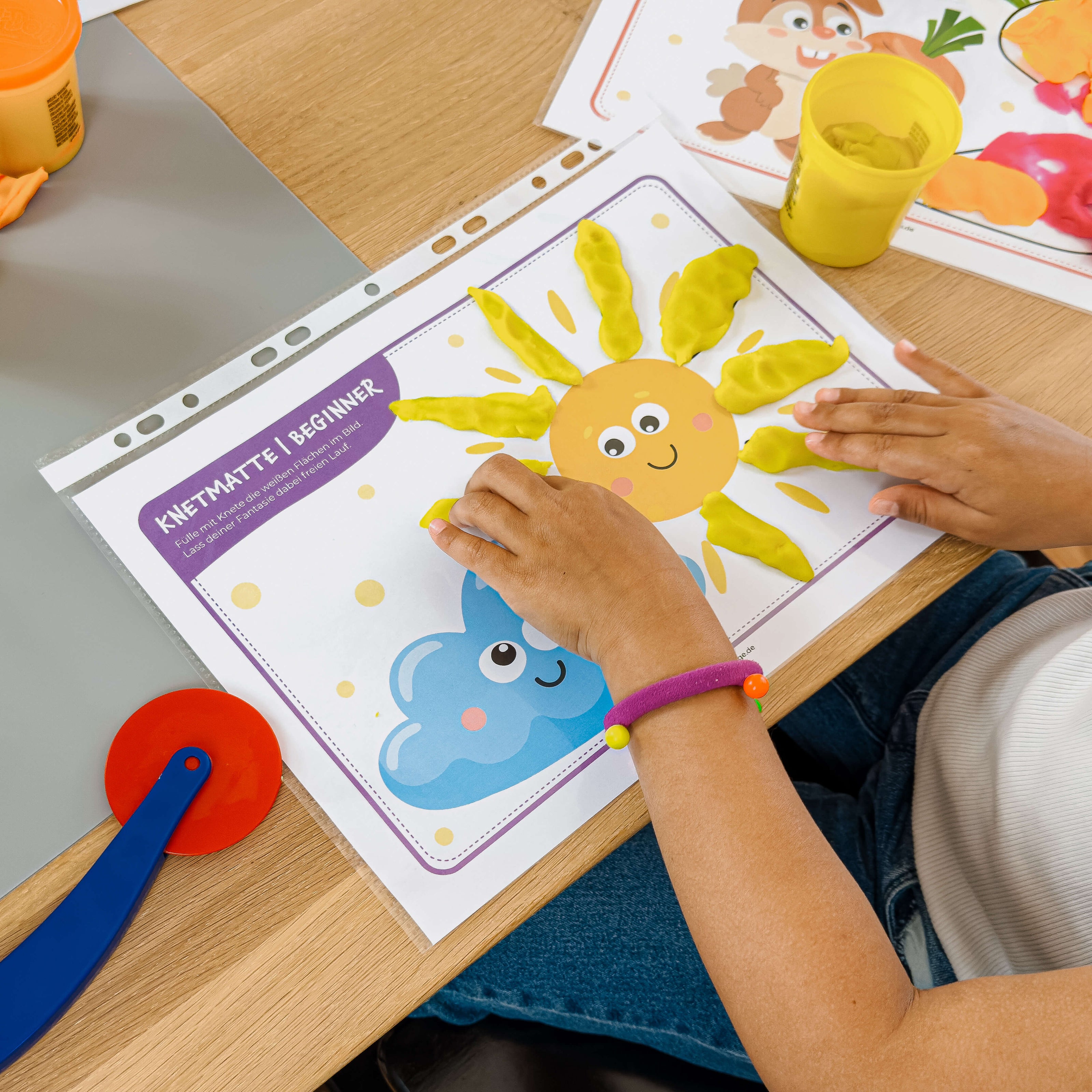Kind legt mit gelber Knete Sonnenstrahlen auf eine fröhliche mykidslounge Beginner Knetmatte mit Sonne und Wolke – spielerisches Training für Fingerkraft und Kreativität.