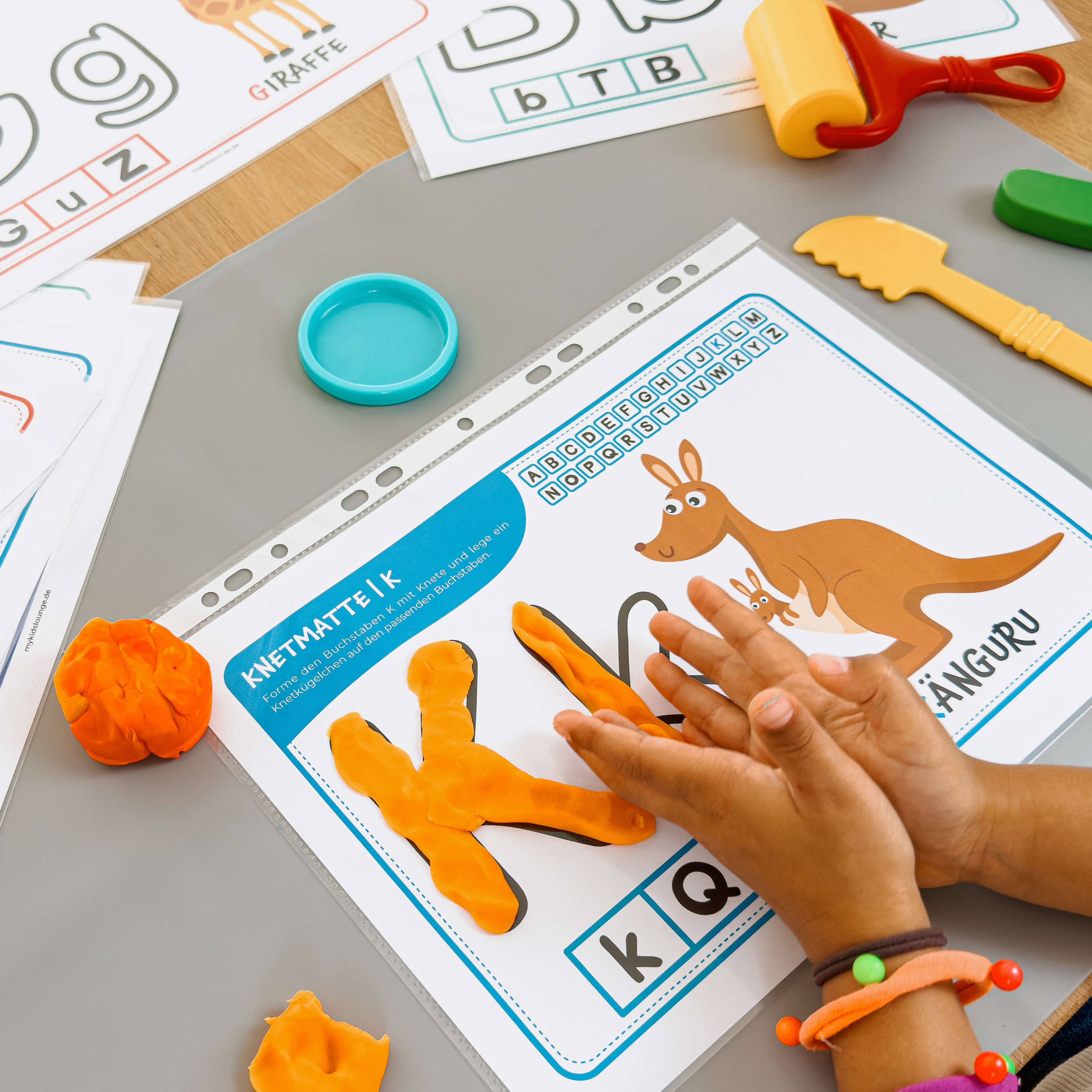 Lernen durch Kneten: Kind gestaltet den Buchstaben K mit oranger Knete auf der Knetmatte mit Känguru von mykidslounge.