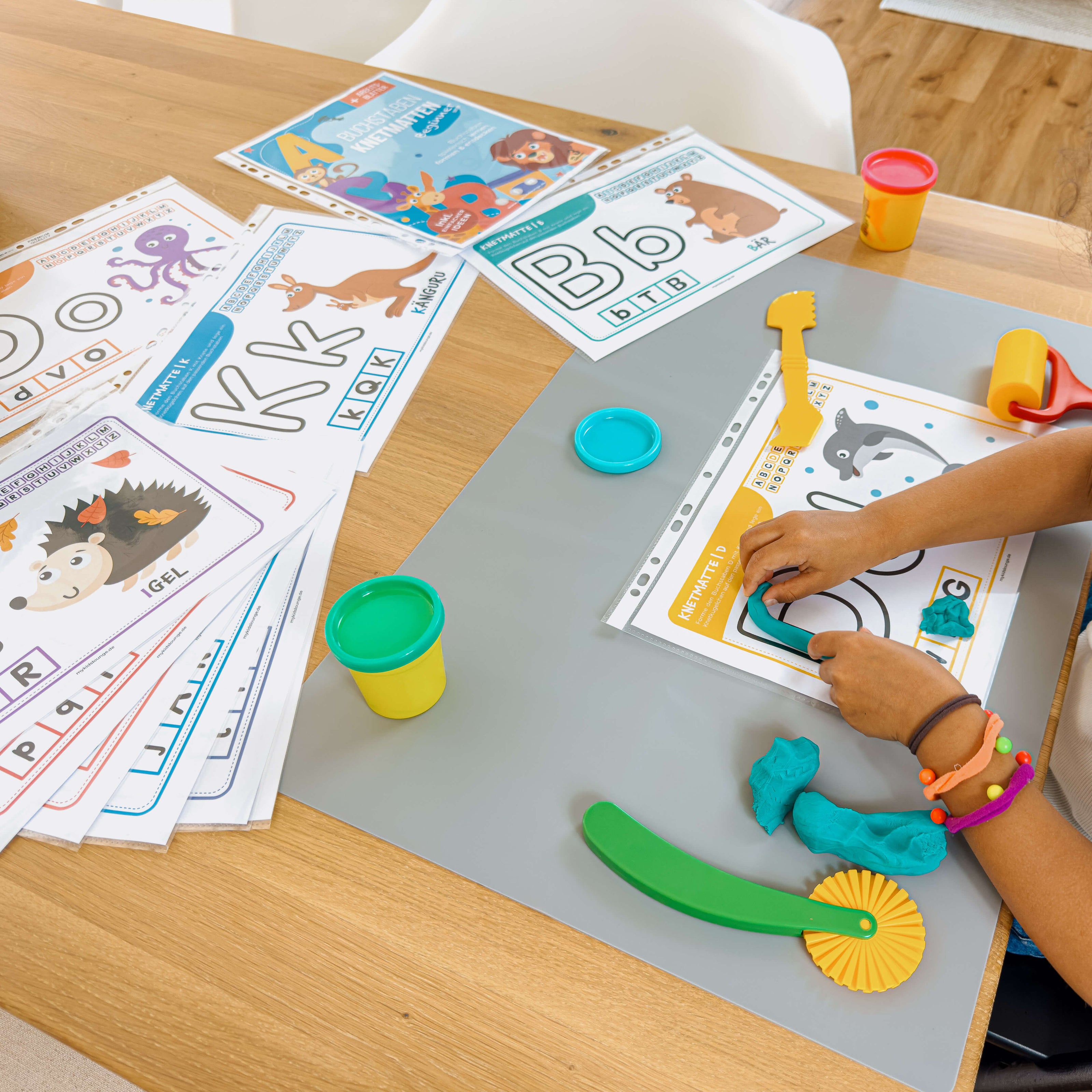 Kind formt den Buchstaben D mit türkisfarbener Knete auf einer Lernmatte von mykidslounge – im Bild liegen weitere liebevoll illustrierte Buchstabenmatten mit Tieren bereit.