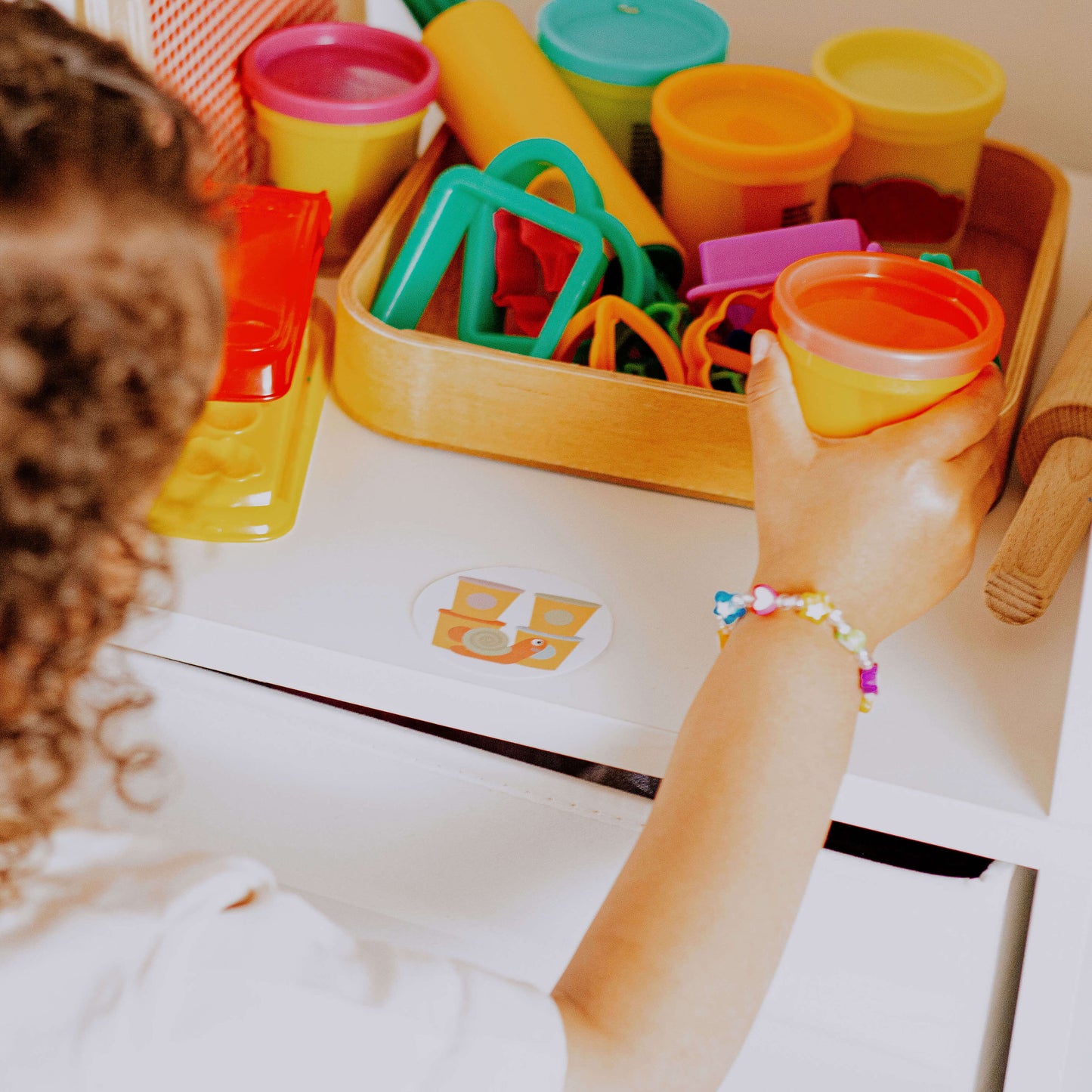 Kleinkind Mädchen räumt Knete und Knetformen zurück ins das Spielzeugregal mit dem Spielzeugetikett für Knete